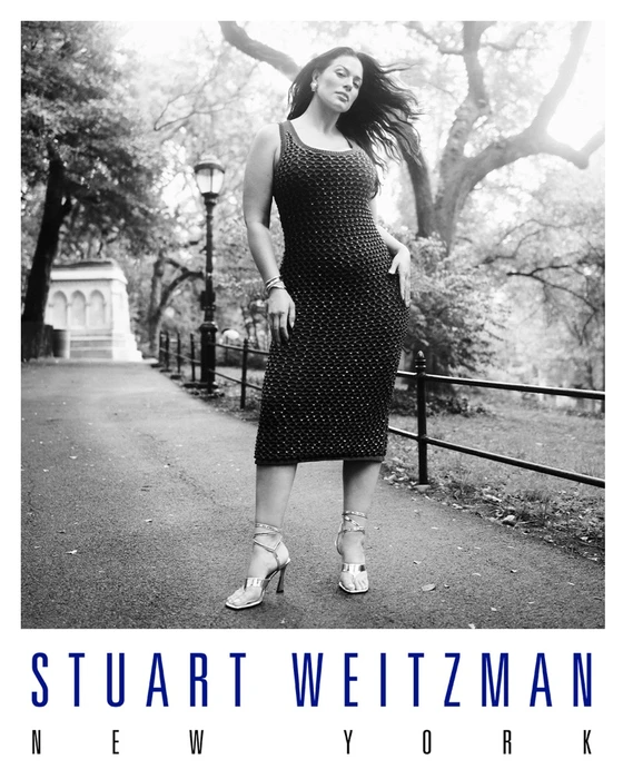 Model Ashley Graham in a dark textured dress and strappy sandals posing on a paved park path for Stuart Weitzman.