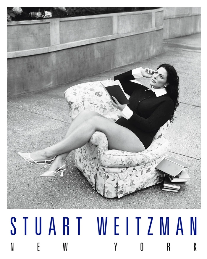 Ashley Graham seated in an armchair outdoors holding a book for the Stuart Weitzman Concrete Paradise campaign.
