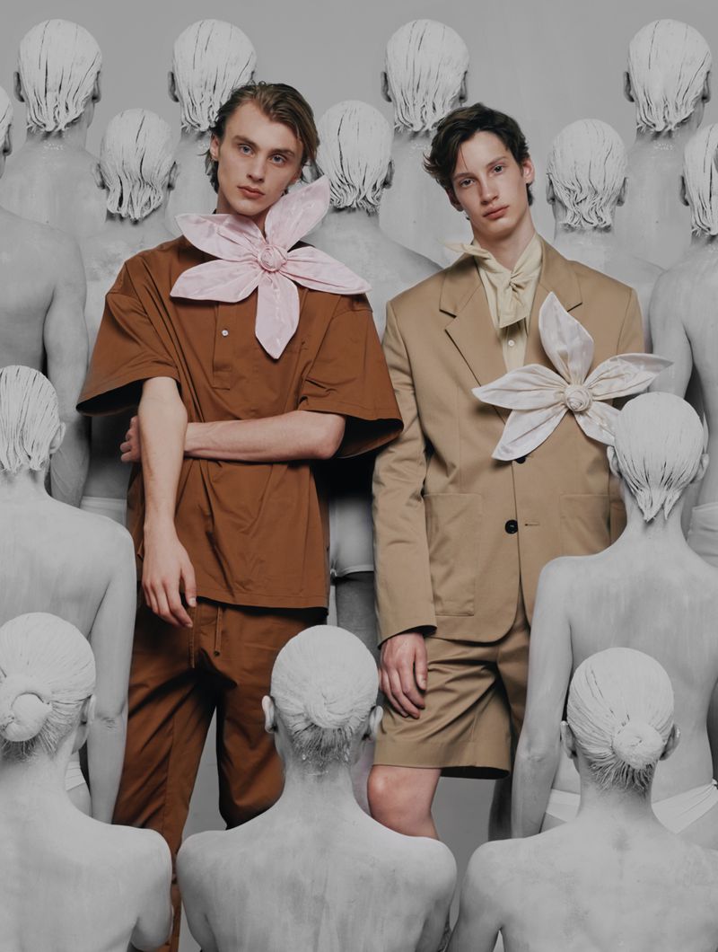 Two young men in jackets and shorts pose in a photo studio with multiple statue figures and neutral lighting, fashion editorial concept.