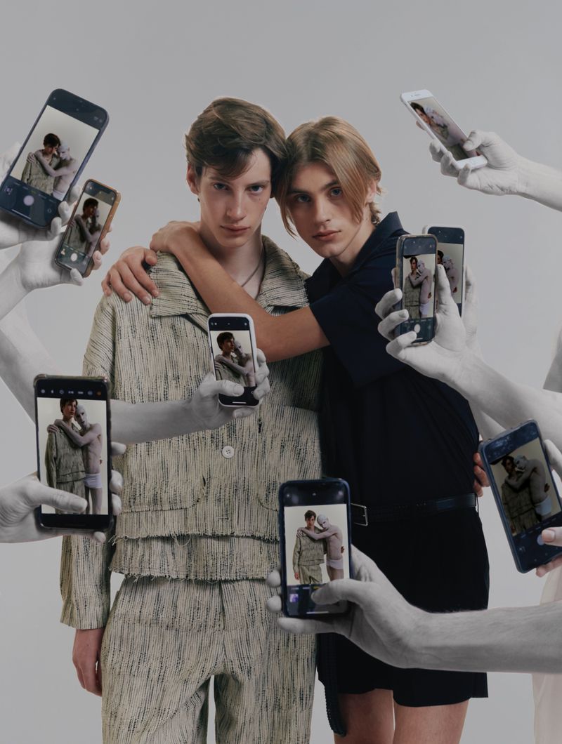 Two young men embrace, phones held up by grayscale hands surround them, capturing the moment in a brightly lit studio setting.