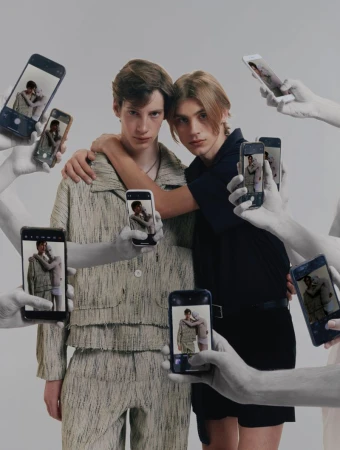 Two young men embrace, phones held up by grayscale hands surround them, capturing the moment in a brightly lit studio setting.