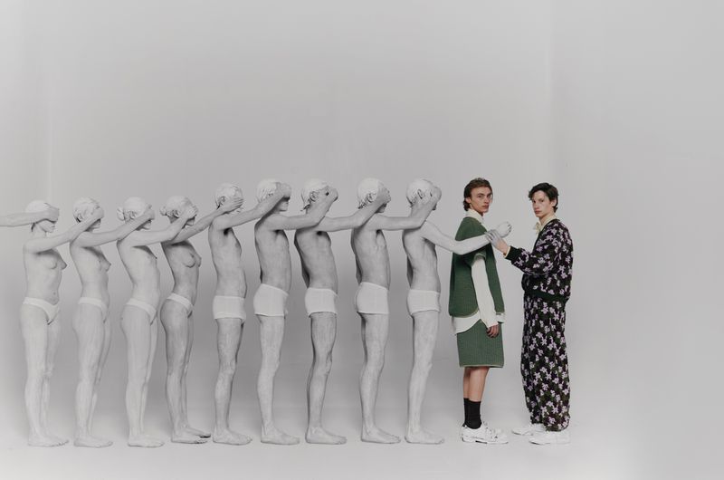 Artistic fashion shot features two women in stylized clothing, contrasted by a line of nude figures with hands over their eyes in a white studio.