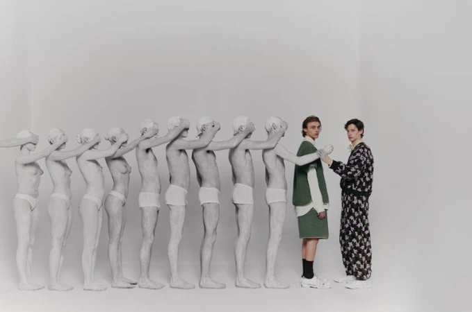Artistic fashion shot features two women in stylized clothing, contrasted by a line of nude figures with hands over their eyes in a white studio.