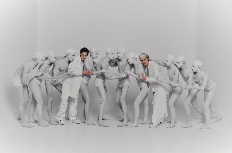 Two adult men in white garments are surrounded by mannequin-like people with white body paint, standing in a bright white studio setting.