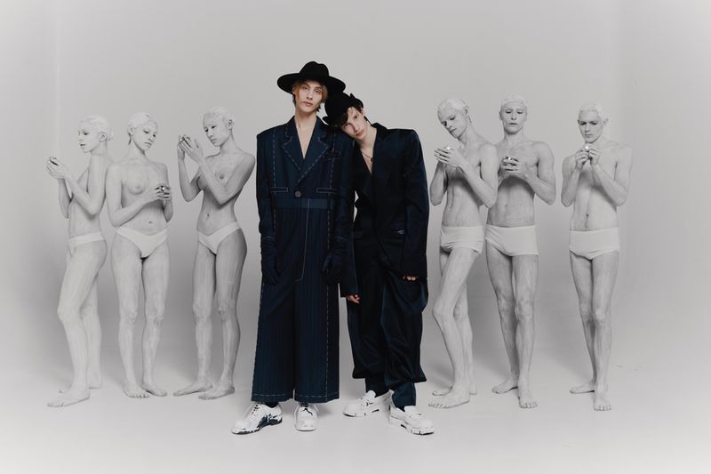 Two models in oversized dark clothing and sneakers pose in a studio with white-painted figures that hold phones. Studio lighting.