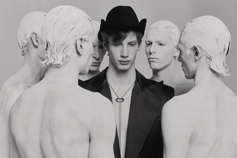 Artistic fashion portrait of a young male model wearing a black jacket and hat, surrounded by five men with white painted skin.