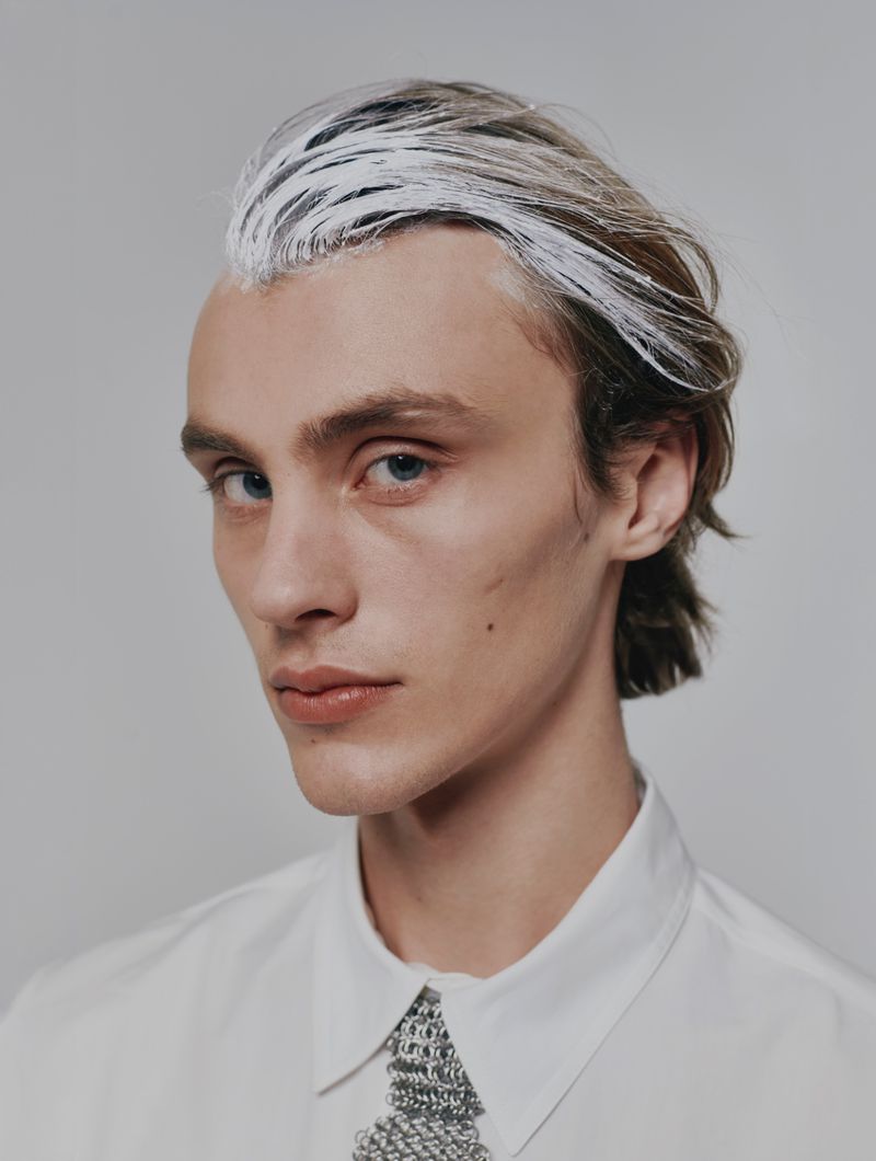 Close-up portrait of a young adult male with blue eyes and light brown hair with white streaks, wearing a white shirt against a gray background.