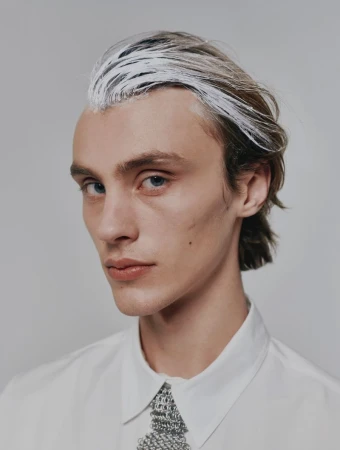 Close-up portrait of a young adult male with blue eyes and light brown hair with white streaks, wearing a white shirt against a gray background.