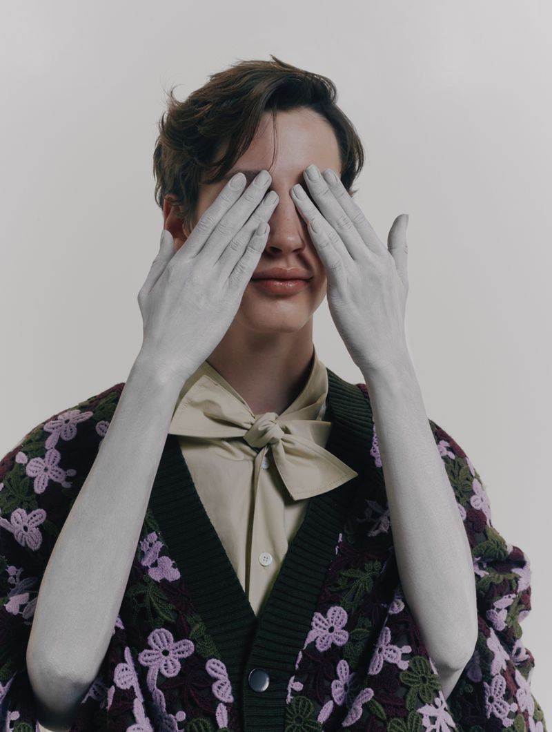 A light-skinned adult with brown hair covers their eyes with silvery hands, wearing a cardigan and bowtie indoors on a neutral background.