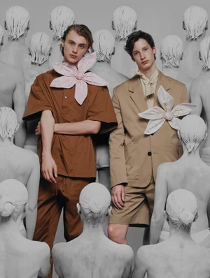 Two male models in brown and tan suits adorned with large fabric flower accessories standing among white statues.