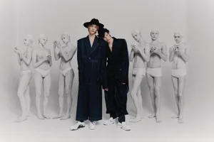 Two central models in dark coats and hats flanked by figures painted white against a seamless grey studio background