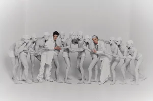 A line of figures painted white interacting in a studio setting with two clothed men standing amongst them.