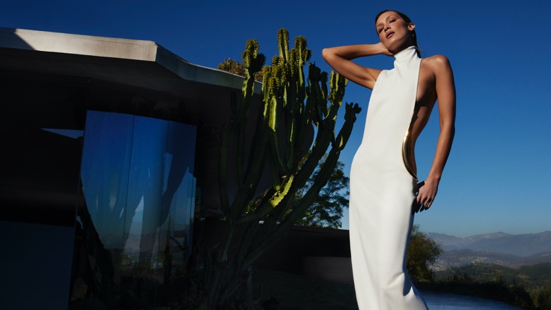 Bella Hadid posing in a high neck white cutout gown with gold hardware beside a cactus under clear blue skies