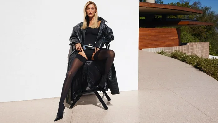 Bella Hadid seated on a director chair wearing a black leather coat and bodysuit against a white and modernist background.