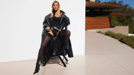 Bella Hadid seated on a director chair wearing a black leather coat and bodysuit against a white and modernist background.