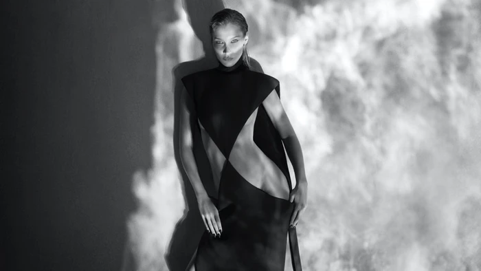 Bella Hadid posing in a dark geometric dress against a projected smoke pattern backdrop in a black and white portrait.