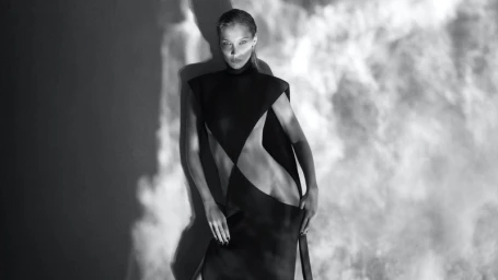 Bella Hadid posing in a dark geometric dress against a projected smoke pattern backdrop in a black and white portrait.