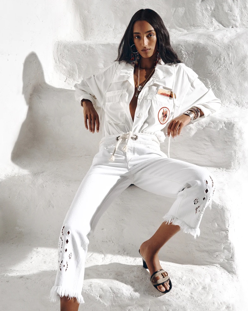 Mona Tougaard seated on whitewashed stone steps wearing a white utility jumpsuit and heeled slides.