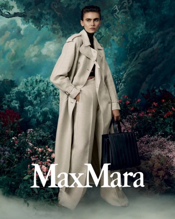 Mia Armstrong wears a beige trench coat and wide-leg trousers holding a black handbag against a painterly floral woodland backdrop.
