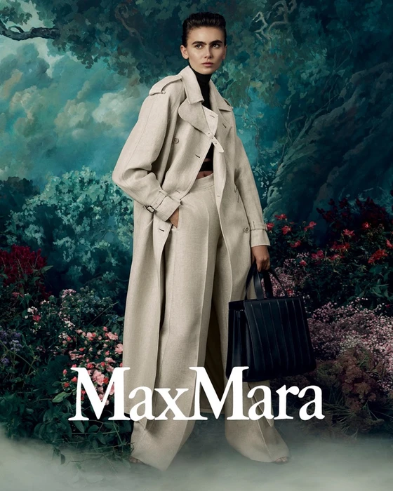 Model wearing a neutral beige trench coat and matching trousers, holding a black handbag against a painterly forest backdrop for Max Mara.