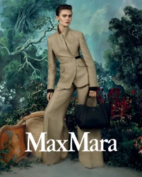 Model standing in a beige tailored pantsuit holding a black leather tote bag against a painted forest backdrop