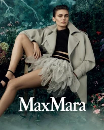 Model seated on a chair wearing a beige trench coat and layered skirt against a lush floral background