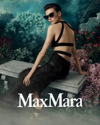 Model wearing dark sunglasses and a black outfit seated on a stone bench surrounded by lush flowering bushes for Max Mara.