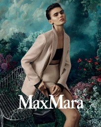 Model Mia Armstrong wearing a beige blazer and shorts holding a black handbag against a painterly floral background for Max Mara.