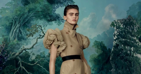 Model wearing a tan trench coat with exaggerated sleeves against a painted forest backdrop