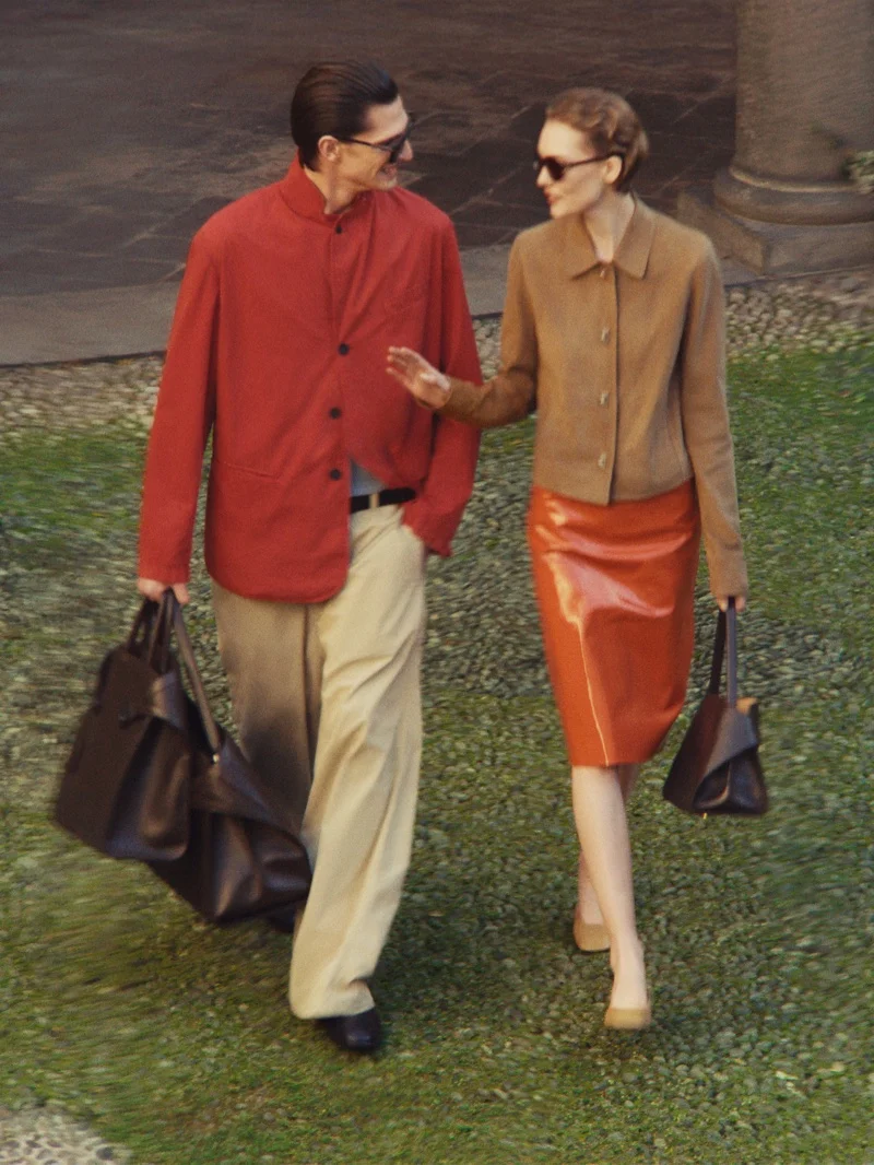 Man in red jacket and woman in brown jacket walking together on stone path holding dark handbags