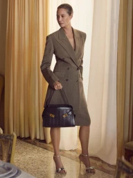 Christy Turlington wearing a tailored brown coat and holding a black leather handbag in a bright hotel room