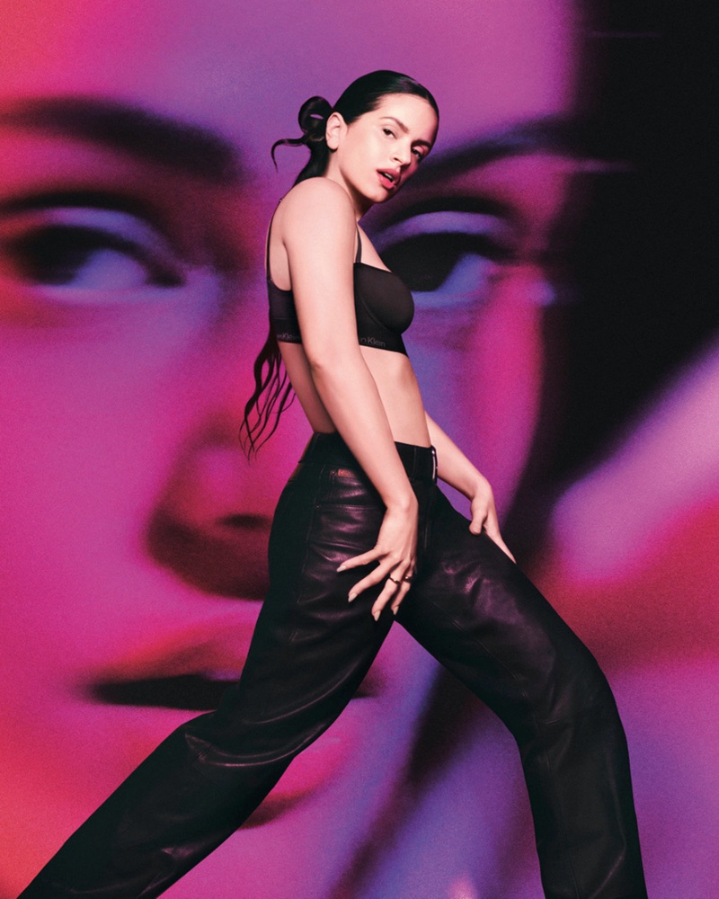 Rosalia posing in a black crop top and leather pants against a vibrant projected face backdrop
