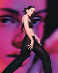 Rosalia posing in a black crop top and leather pants against a vibrant projected face backdrop