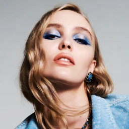 Portrait of Lily-Rose Depp wearing blue eye makeup and a denim jacket against a neutral grey background.