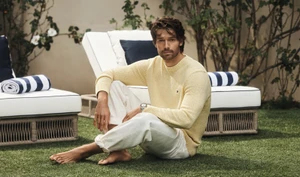 Patrick Schwarzenegger seated on grass in a yellow sweater and white trousers for Tommy Hilfiger