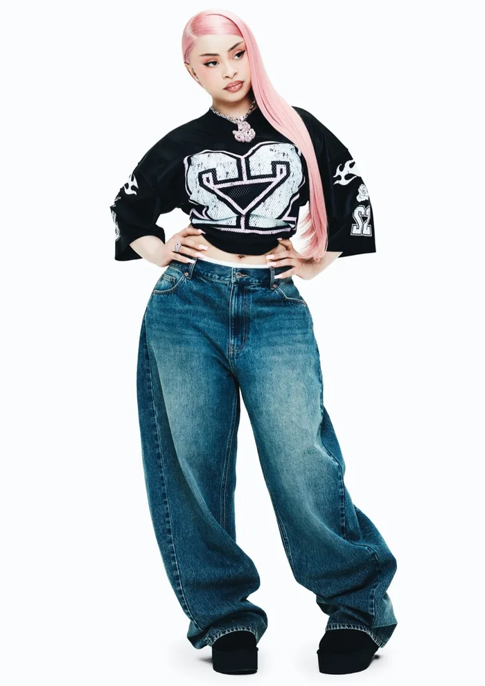 Ice Spice with pink hair wearing a black graphic t-shirt and baggy denim jeans against a white background