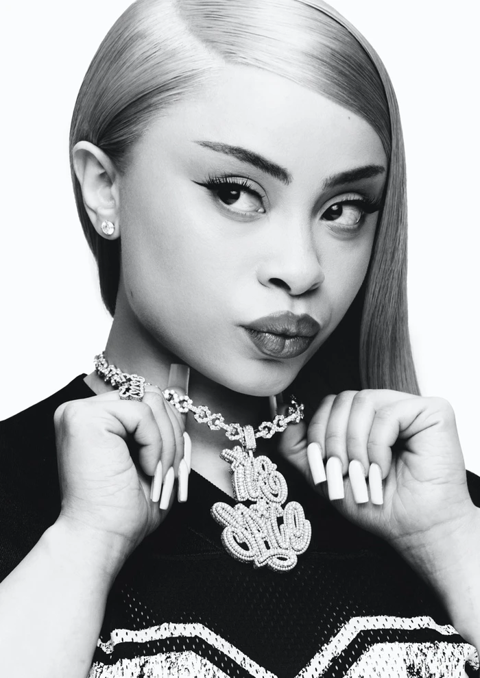 Portrait of Ice Spice wearing a large diamond pendant necklace and dark top against a plain white background.
