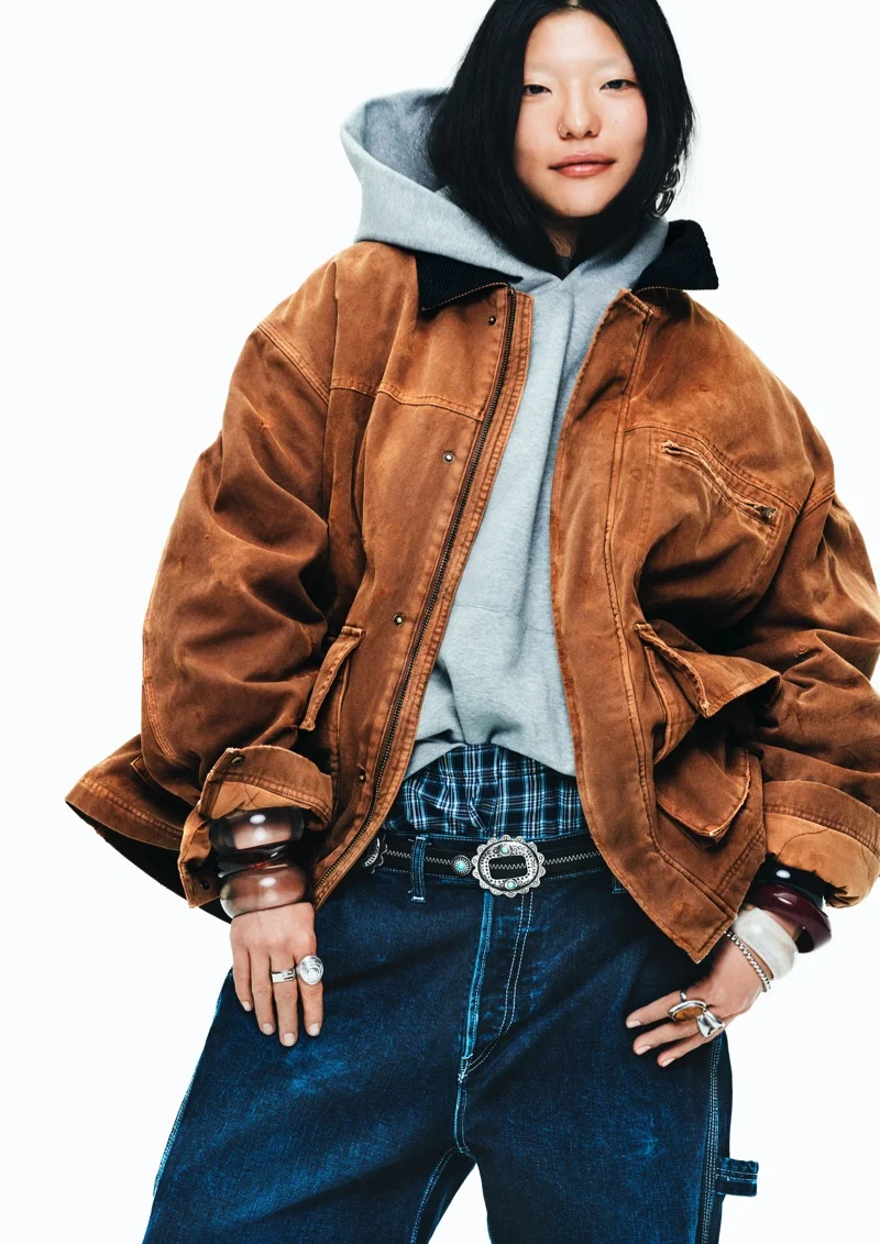 Model wearing a brown jacket over a grey hoodie and blue jeans against a white backdrop