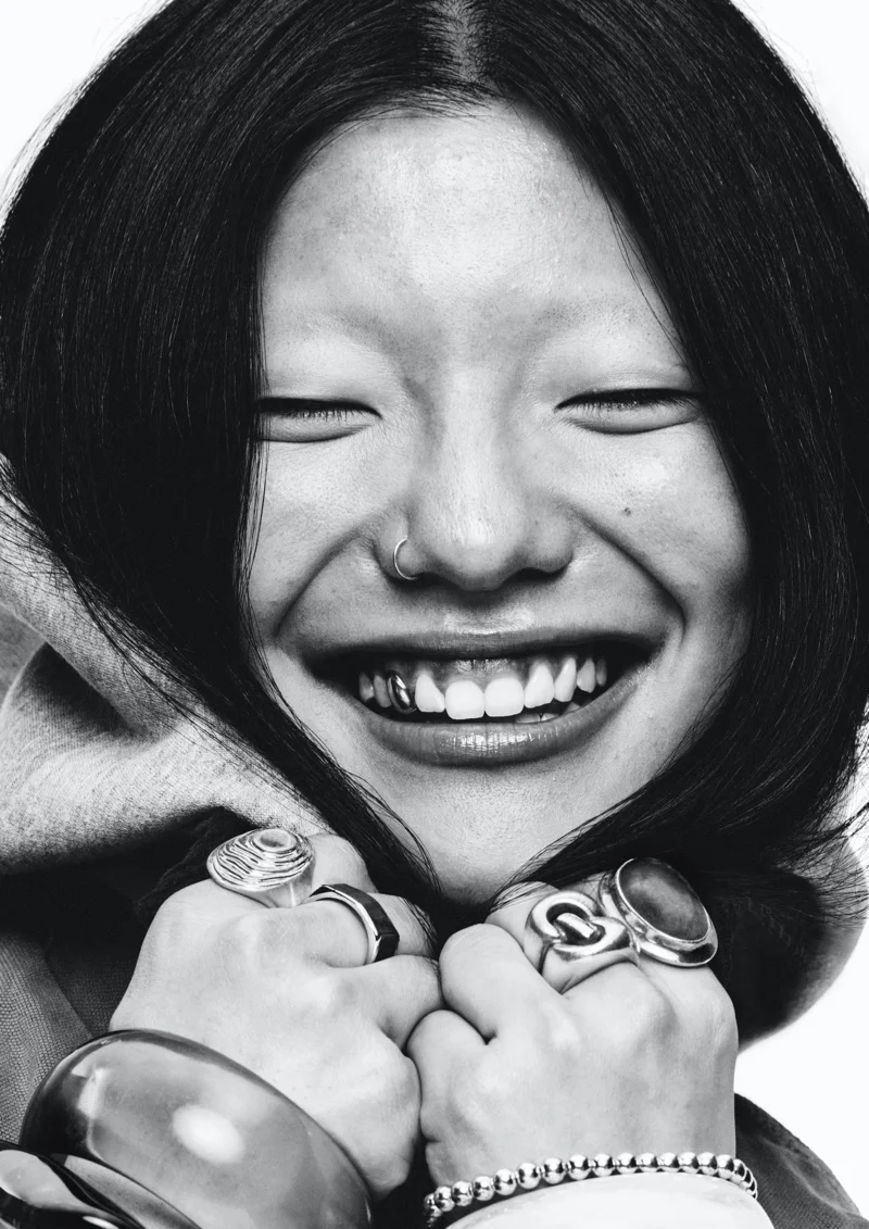Close-up monochrome portrait of a smiling person with dark hair, nose ring, and multiple rings on hands against white background.