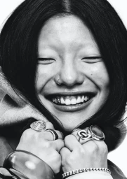 Close-up monochrome portrait of a smiling person with dark hair, nose ring, and multiple rings on hands against white background.