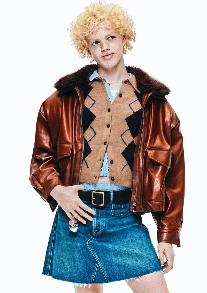 Model with curly blonde hair wearing a brown leather jacket, argyle cardigan, and denim skirt against white background.