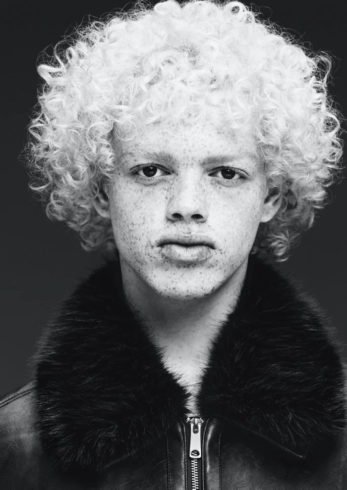 Portrait of a young man with curly blonde hair and freckles wearing a black jacket with a fur collar