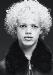 Portrait of a young man with curly blonde hair and freckles wearing a black jacket with a fur collar