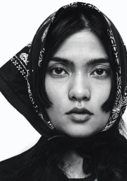 Close-up portrait of a woman wearing a black patterned headscarf and a septum nose ring against a white background.