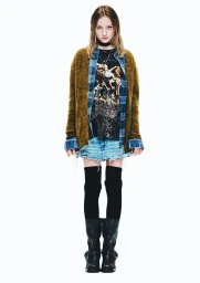 Full-length portrait of a female model wearing an olive cardigan, blue plaid shirt, graphic tee, distressed denim skirt, and black boots against a white background.