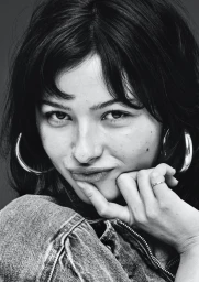Close-up black and white portrait of a woman wearing hoop earrings and denim jacket, resting chin on finger