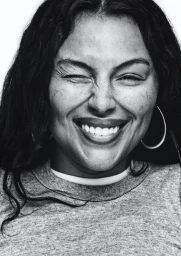 Close-up portrait of Paloma Elsesser winking and smiling broadly, wearing a crewneck t-shirt and hoop earrings against a white background.