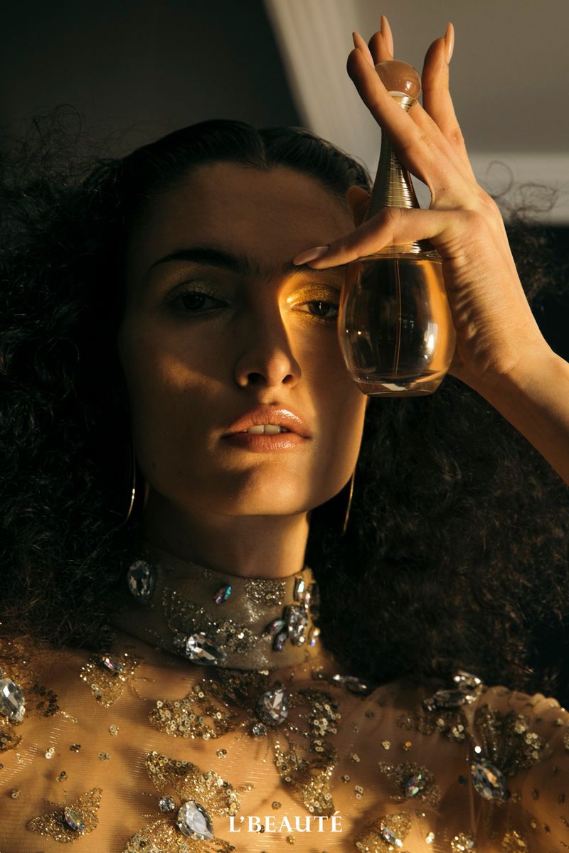 Beauty shot of woman holding gold perfume bottle in front of eye wearing an ornate jeweled dress, dark curly hair, and gold eye makeup.