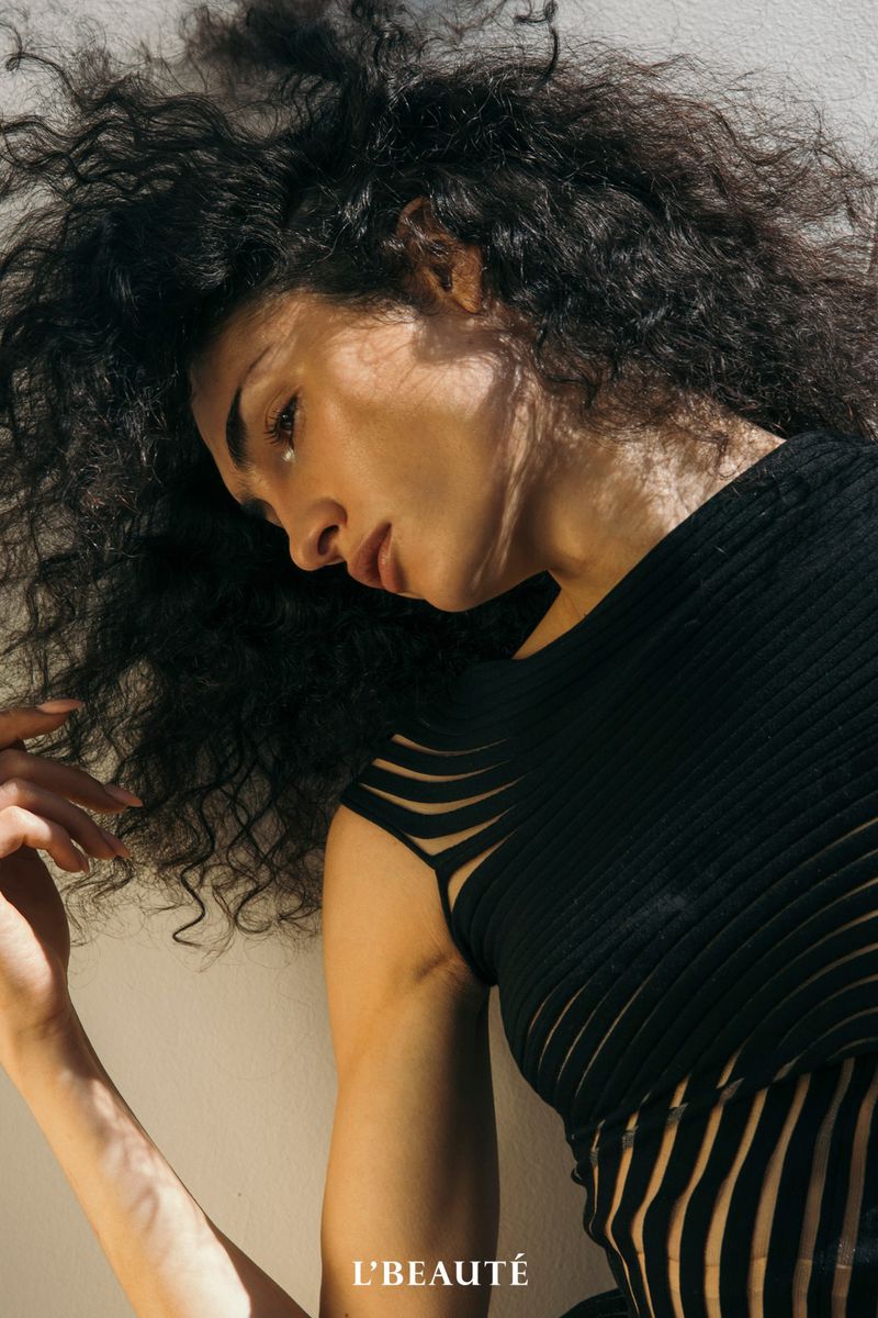Portrait of a woman with curly hair wearing a black top, looking down. The image features natural light and a minimalist backdrop.