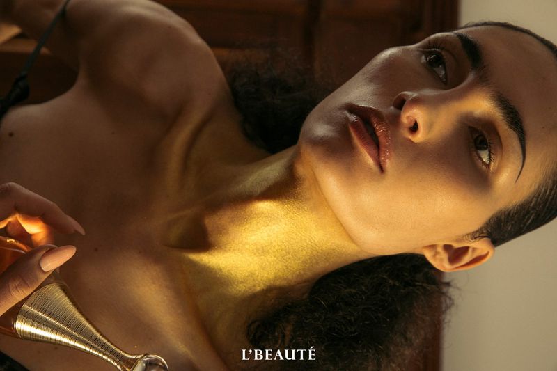 Adult female lying down with a golden, glossy skin tone. She is holding a perfume bottle in soft, diffused lighting. Beauty shot.